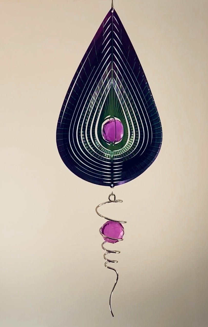 Purple teardrop wind spinner with layered purple and green rings, magenta crystal centre and silver spiral crystal tail, hanging against a beige wall